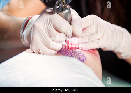 Close-up of a young woman tattoo master in sterile gloves makes a tattoo on the thigh with red paints Stock Photo