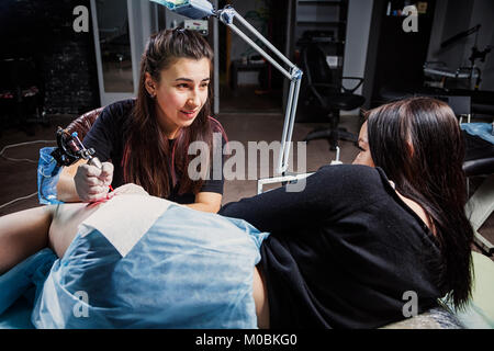 A young woman tattoo artist doing a tattoo with a tattoo machine under the bright light of a lamp on a beautiful woman with whom she talks in a dark w Stock Photo