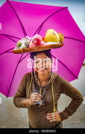 Filipino woman with a basket of fruit on her head and a bright, magenta colored umbrella smiles at the camera in Baloy Long Beach, Luzon, Philippines. Stock Photo
