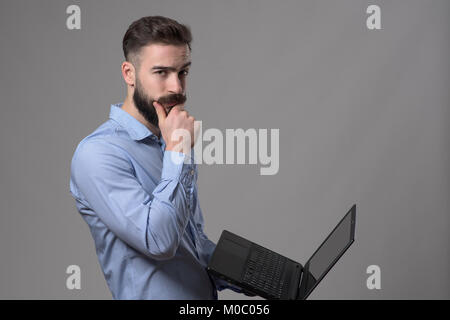 Confused young business man using laptop computer touching beard and looking at camera against gray studio background. Stock Photo