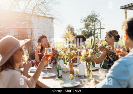 Group of young friends hanging out with drinks at outdoors party. Young men and women sitting around a table toasting wine. Stock Photo