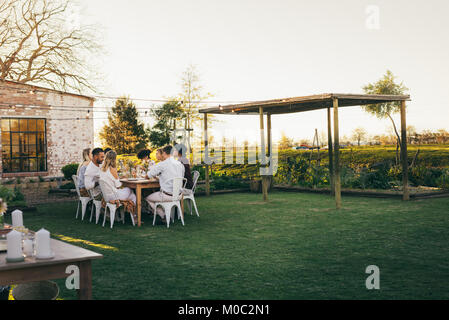 Group of young friends sitting around a table in garden restaurant and having meal. Young men and women having outdoors party. Stock Photo