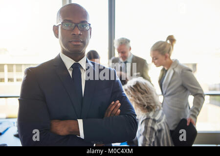 Portrait of a focused young businessman in glasses standing in a bright modern office with a group of colleagues working in the background Stock Photo