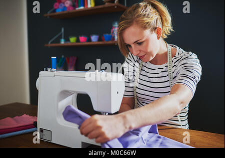 Young creative fashion designer using a sewing machine to stitch a purple fabric in the interior of a workshop Stock Photo