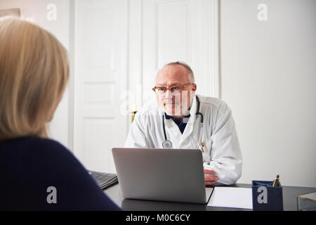 Mature doctor sitting and smiling at laptop in office and talking to female patient. Stock Photo