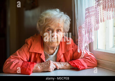 Portrait of an elderly woman sitting in the cloak at the table. Stock Photo