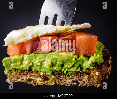 Bite sized piece of avocado toast with tomato and a fried egg on a fork.  Isolated on a black background Stock Photo