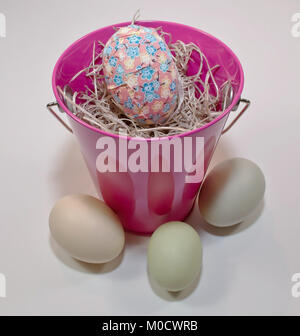 A single Easter egg in a bed of straw within a pink bucket, three other eggs are in front of the bucket on a white background Stock Photo