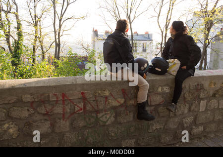 Two motorcyclists chatting while sitting on a stone wall looking out over Paris in Montmartre, France with motorbike helmets between them Stock Photo