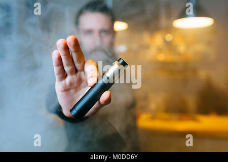 Young beard man show vaping device on his outstretched hand through a cloud of steam. Selective focus. Vaping concept. Copy space. Stock Photo