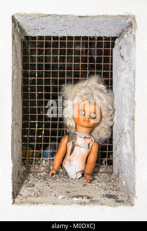 Spooky scene with a broken abandoned doll. Old damaged toy in window with rusty metal grid. Concept of science fiction, apocalypse, war or abuse. Stock Photo