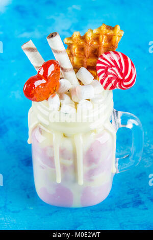 Freakshake from pink smoothie, cream. Monstershake with lollipops, waffles and marshmallow. Extreme milkshake in a Mason jar. Blue concrete background Stock Photo