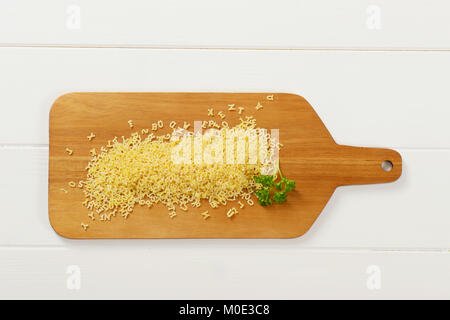 heap of uncooked alphabet pasta on wooden cutting board Stock Photo