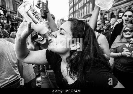 London black and white street photography: Young woman cheered on by crowd as she drinks from bottle at street party. Stock Photo