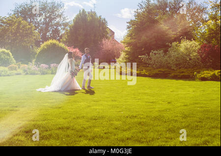 Beautiful just married couple walking in meadow full of sunset warm light with trees and building on background Stock Photo