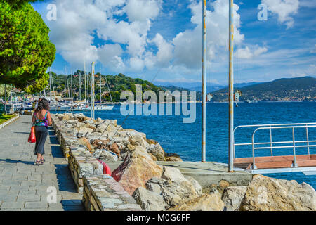 A woman walks along the boardwalk at the port harbor of Portovenere Italy on a sunny day in the Gulf of Poets with sailboats and mountains behind Stock Photo