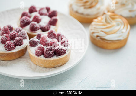 Delicious lemon and raspberry tartlets with meringue on a white vintage plate. Sweet treat on a light blue background. Stock Photo