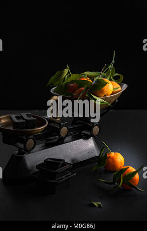 A bunch of fresh juicy clementines with green leaves in a scalepan and on a black table Stock Photo
