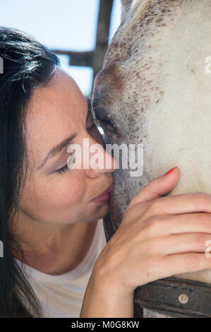 black haired female cuddling and kissing a white horse in cheek, close up Stock Photo
