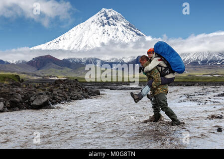 Guide helps young women tourist to cross muddy rugged mountain river - strong man carries woman on her back on background beautiful volcano cone. Stock Photo