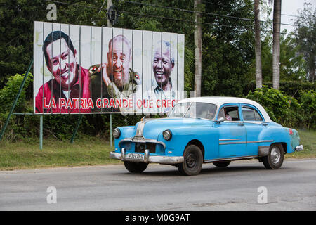 Classic American car driving past billboard of Hugo Chavez, Fidel Castro and Nelson Mandella in Cienfuegos, Cuba Stock Photo