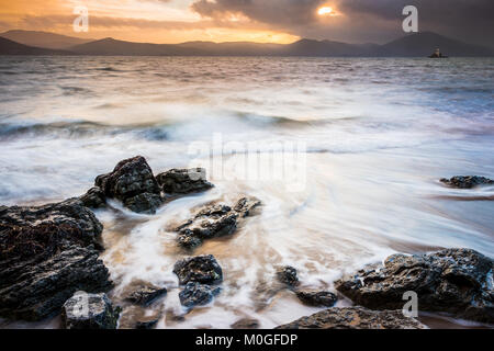 Waves recede during a stormy sunset. A lighthouse and mountains can be seen in the background. Stock Photo