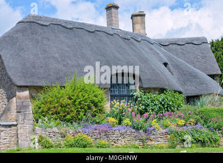 Old traditional English limestone cottage with thatched roof, big window, colourful flowering front garden, on a summer sunny day in Cotswolds, United Stock Photo