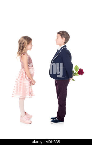 side view of stylish boy in suit holding rose flower and looking at beautiful little girl in pink dress isolated on white  Stock Photo