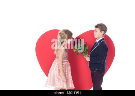 side view of little boy presenting flowers to beautiful little girl and big red heart symbol behind isolated on white  Stock Photo