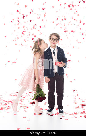 cute little boy in eyeglasses and suit holding gift box and smiling at camera while girl with roses able to kiss him on white with falling heart shaped confetti Stock Photo