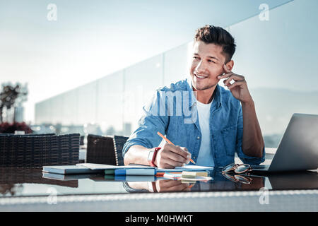 Pensive young gentleman smiling while working on laptop outdoors Stock Photo