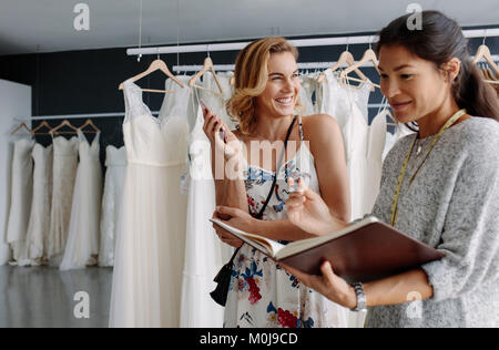 Female tailor making notes in her diary with bride in wedding fashion store. Wedding dress designer taking order from female client in bridal wear sho Stock Photo