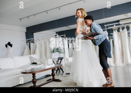 Female trying on wedding gown with women assistant in bridal wear shop. Smiling woman wearing her bridal dress with wedding dress designer in bridal f Stock Photo