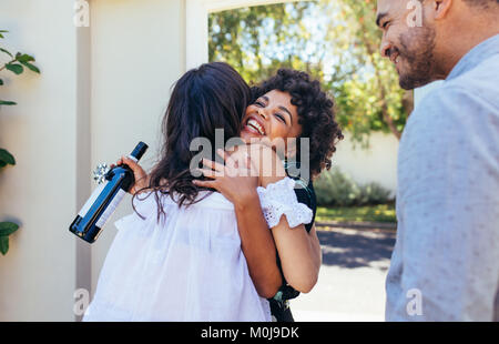 Woman greeting couple for having a new house. Smiling young woman with wine bottle congratulating her friend. Housewarming party with friends. Stock Photo