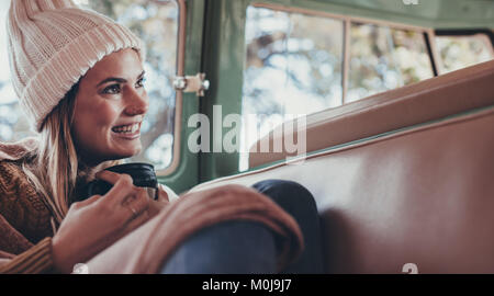 Woman on roadtrip travelling by van. Smiling woman in warm clothing  sitting in back seat of van with cup of coffee. Stock Photo