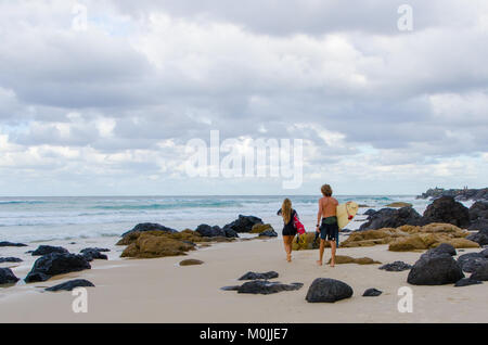Rear view of male and female surfers walking across a rocky beach towards the sea - Kirra Beach, Coolangatta, Gold Coast, Australia