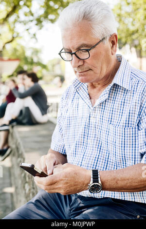 Grey-haired senior sits on a wall with his smartphone in his hand, Cologne, North Rhine-Westphalia, Germany Stock Photo