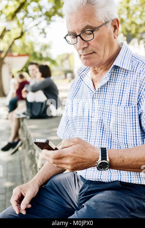 Grey-haired senior sits on a wall with his smartphone in his hand, Cologne, North Rhine-Westphalia, Germany Stock Photo