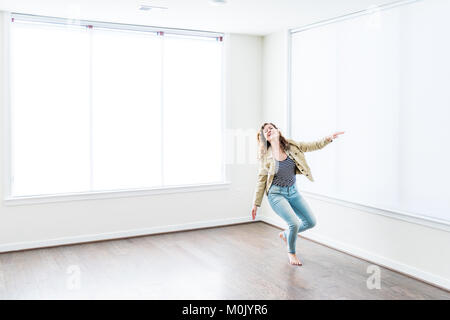 One young happy smiling woman jumping up in empty modern new room with hardwood floors and large sunny windows in apartment Stock Photo