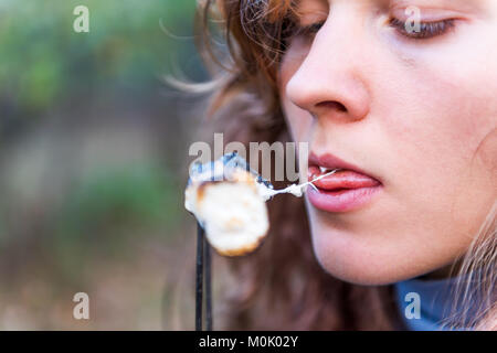 One young woman eating roasted caramelized marshmallow skewer closeup macro portrait showing teeth, biting, mouth, face, tongue Stock Photo