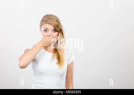 Saying bad things, shocking news concept. Ashamed young blonde woman having hand on her mouth. Studio shot on grey background. Stock Photo