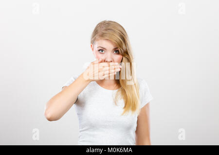 Saying bad things, shocking news concept. Ashamed young blonde woman having hand on her mouth. Studio shot on grey background. Stock Photo