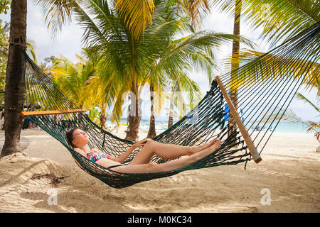 A young woman in a bikini lays in a hammock on a tropical beach with the ocean in the background; Negril, Jamaica Stock Photo