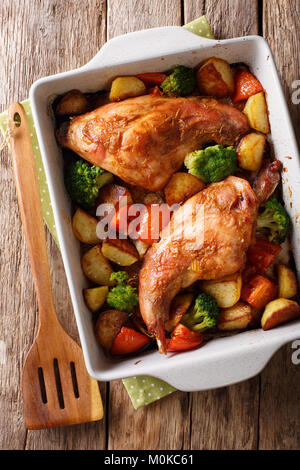 Delicious baked rabbit legs with potatoes, broccoli and tomatoes close-up in a baking dish on a table. Vertical top view from above Stock Photo