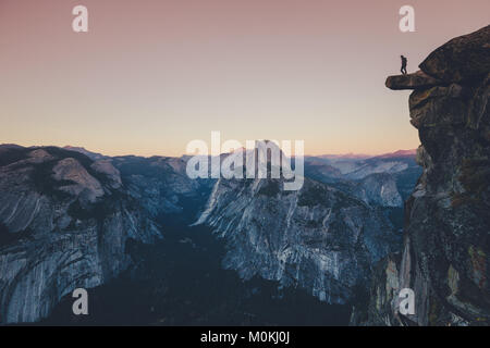 A fearless hiker is standing on an overhanging rock looking towards famous Half Dome in Yosemite National Park in twilight at sunset, California, USA Stock Photo