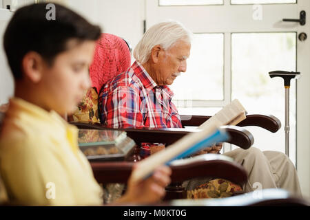 Grandson And Grandpa Reading Book And Relaxing At Home Stock Photo