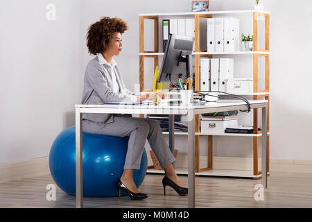 Young Businesswoman Using Computer While Sitting On Fitness Ball In Office Stock Photo