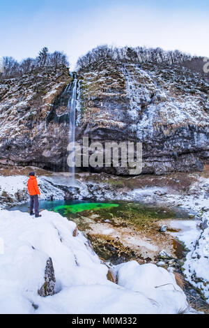 Man in orange jacket standing near waterfall Goriuda in winter wonderland in Italian alps Stock Photo
