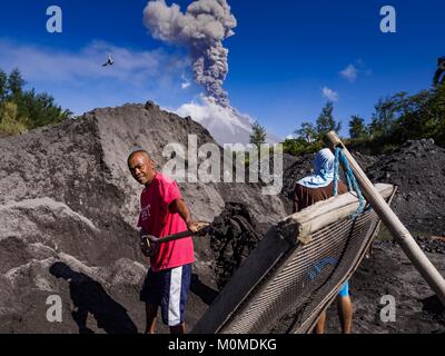 Daraga, Albay, Philippines. 23rd Jan, 2018. Sand and gravel miners work in a riverbed of volcanic soil while the Mayon volcano erupts Tuesday. The Mayon volcano continued to erupt Tuesday, although it was not as active as it was Monday. There were ash falls in communities near the volcano. This is the most active the volcano has been since 2009. Schools in the vicinity of the volcano have been closed and people living in areas affected by ash falls are encouraged to stay indoors, wear a mask and not participate in strenuous activities. Credit: Jack Kurtz/ZUMA Wire/Alamy Live News Stock Photo