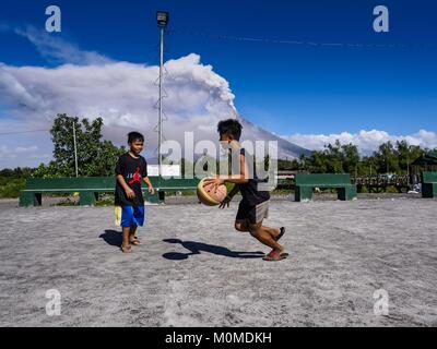 Daraga, Albay, Philippines. 23rd Jan, 2018. Children play basketball on a recreation yard in Daraga while the Mayon volcano erupts Tuesday. The Mayon volcano continued to erupt Tuesday, although it was not as active as it was Monday. There were ash falls in communities near the volcano. This is the most active the volcano has been since 2009. Schools in the vicinity of the volcano have been closed and people living in areas affected by ash falls are encouraged to stay indoors, wear a mask and not participate in strenuous activities. Credit: Jack Kurtz/ZUMA Wire/Alamy Live News Stock Photo